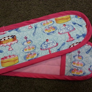 Puede incluir: Un par de guantes de horno rosas con un patrón azul y blanco de pasteles y postres. Los guantes están acolchados y tienen un ribete rosa.