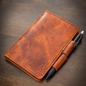 May include: A brown leather notebook with a pen tucked into the cover. The notebook has a stitched edge and a worn, distressed look.