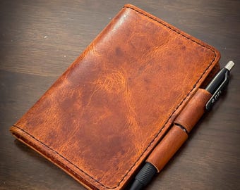 May include: A brown leather notebook with a pen tucked into the cover. The notebook has a stitched edge and a worn, distressed look.