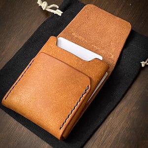 May include: A handcrafted, open leather card holder in a warm tan color, with visible stitching and a contrasting dark blue thread. The card holder is partially open, revealing white cards inside. It rests on a black drawstring bag, set against a dark wood surface.