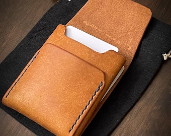 May include: A handcrafted, open leather card holder in a warm tan color, with visible stitching and a contrasting dark blue thread. The card holder is partially open, revealing white cards inside. It rests on a black drawstring bag, set against a dark wood surface.