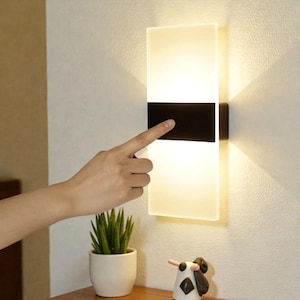 Puede incluir: Una lámpara de pared moderna con una pantalla rectangular de vidrio esmerilado, dividida por una banda negra. La luz está encendida y un dedo toca la sección negra. Una pequeña planta en maceta y una figura decorativa están en primer plano.