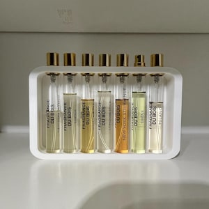 May include: A white tray contains ten small perfume bottles with gold lids. Each bottle is labelled "FRAGRANCE DU BOIS" with various fragrance names. The liquids inside range in colour from clear to amber and yellow.