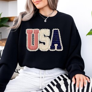 May include: Black sweatshirt with the word "USA" in a patriotic design. The letters are in red and white stripes, beige, and navy blue with white stars. The letters are outlined with a white lace trim.