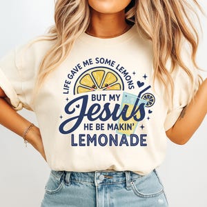 May include: Cream-colored t-shirt with a graphic design. The design features a smiling lemon slice, a glass of lemonade, and the text "Life gave me some lemons but my Jesus he be makin' lemonade" in navy blue.
