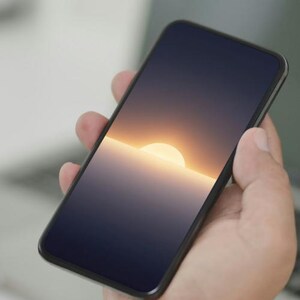 May include: A black smartphone held in a hand, displaying a sunset over the ocean. The screen shows a gradient of colors, with a bright yellow sun setting over a dark blue sea. The phone has a sleek, modern design.