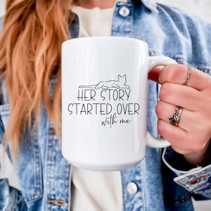 Peut inclure: Mug en céramique blanche avec un dessin au trait noir d'un chat et le texte "HER STORY STARTED OVER with me". La tasse a une grande anse. Une veste en jean est visible.