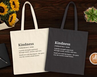 Peut inclure: Deux sacs fourre-tout, un crème et un noir, avec la définition de "Kindness" imprimée en blanc. Les sacs sont sur une surface en bois foncé, avec des tournesols, un carnet, un stylo et une tasse de café.