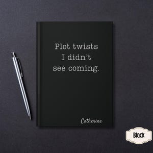 May include: A black notebook with the text "Plot twists I didn't see coming" and the name "Catherine" in white. A silver pen rests on the dark surface next to the notebook. A small "Black" label is in the bottom right corner.