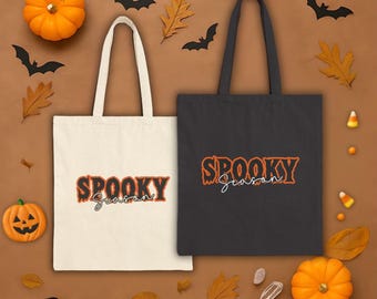 May include: Two tote bags, one cream and one black, with the words "Spooky Season" in orange and white. The bags are surrounded by Halloween decorations, including pumpkins, bats, and autumn leaves.