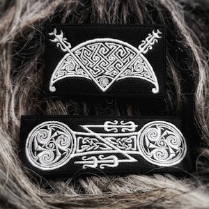 Op de afbeelding: Twee zwarte geborduurde patches met witte Keltische knoopontwerpen. De bovenste patch heeft een halvemaanvorm met twee pijlen die bovenaan elkaar kruisen. De onderste patch heeft een gestileerd knoopontwerp met twee cirkels aan weerszijden.