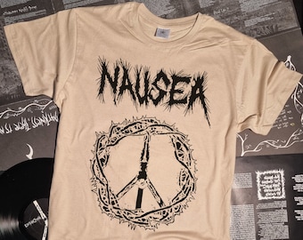May include: Tan-colored t-shirt with the word "NAUSEA" in a spiky font above a peace sign encircled by thorns. The shirt is displayed on a surface with vinyl records and printed materials.