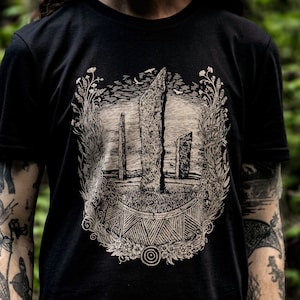 Puede incluir: Camiseta negra con un estampado beige de un círculo de piedras con un diseño floral. El estampado presenta una piedra central con un patrón geométrico.