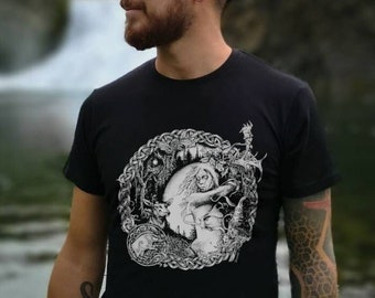 May include: A man wearing a black t-shirt with a white graphic print of a woman with a wolf's head. The graphic is surrounded by a circular design of vines and branches. The man is standing in front of a waterfall.