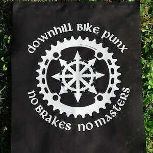 Op de afbeelding: Zwarte vlag met witte tekst en een wit tandwielontwerp. De tekst luidt "downhill bike punx no brakes no masters".