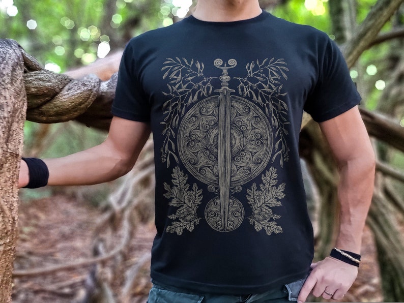 May include: A black t-shirt with a graphic design of a sword and shield surrounded by stylized leaves and berries.