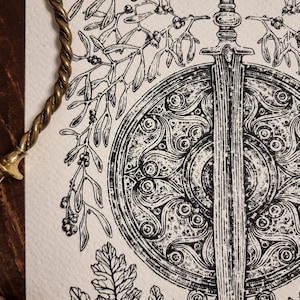 May include: A black and white illustration of a sword and shield with intricate designs. The sword is surrounded by a wreath of mistletoe and oak leaves.