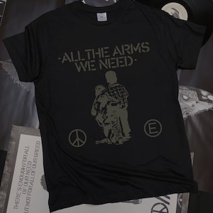 May include: Black t-shirt with a green print of two figures walking. The text "ALL THE ARMS WE NEED." is printed above the figures. A peace sign and a letter "E" are printed on either side of the figures.