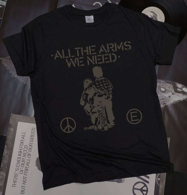 May include: Black t-shirt with a green print of two figures walking. The text "ALL THE ARMS WE NEED." is printed above the figures. A peace sign and a letter "E" are printed on either side of the figures.