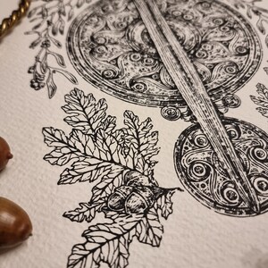 May include: Black and white ink drawing of a sword and shield with intricate Celtic knotwork designs. The sword is positioned vertically, with the shield behind it. A branch with leaves and acorns is in the foreground.