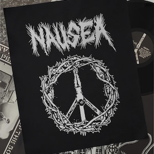 May include: A black fabric patch featuring the white text NAUSEA in a jagged, stylized font at the top. Below the text is a white graphic of a peace symbol encircled by a crown of thorns, with a human figure hanging from the center vertical line.