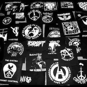 Pode incluir: Uma coleção de patches pretos e brancos com vários desenhos e textos. Alguns dos patches apresentam símbolos anarquistas, sinais de paz e lemas como "No Doves Fly Here", "The System", "Behind Enemy", "Poison Girl" e "Crucify".