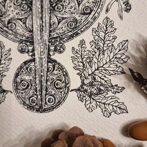 May include: A black and white illustration of a Celtic knot design with a sword in the center. The design is surrounded by leaves and acorns.