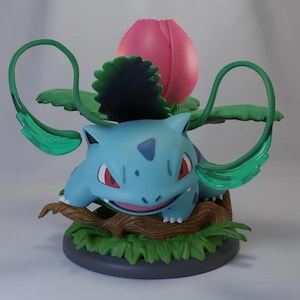 Puede incluir: Una figura azul de Bulbasaur con ojos rojos y una expresión sonriente. La figura presenta una flor rosa y hojas verdes en la espalda, y está colocada sobre una base con hierba y un tronco.