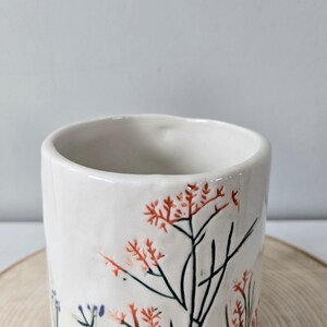 K&ouml;nnte beinhalten: Eine wei&szlig;e Keramiktasse mit floralem Design. Die Tasse hat eine zylindrische Form mit einem leicht ausgestellten Rand. Das florale Design umfasst rote und schwarze Zweige mit kleinen roten Bl&uuml;ten. Die Tasse steht auf einer Holzoberfl&auml;che.