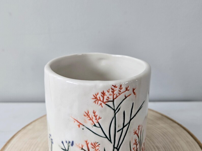 K&ouml;nnte beinhalten: Eine wei&szlig;e Keramiktasse mit floralem Design. Die Tasse hat eine zylindrische Form mit einem leicht ausgestellten Rand. Das florale Design umfasst rote und schwarze Zweige mit kleinen roten Bl&uuml;ten. Die Tasse steht auf einer Holzoberfl&auml;che.
