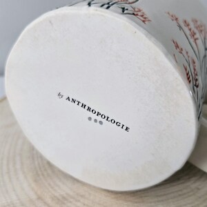 K&ouml;nnte beinhalten: Wei&szlig;e Keramiktasse mit floralem Design in Rot und Schwarz. Die Tasse hat einen gebogenen Henkel und den Text "by ANTHROPOLOGIE" auf der Unterseite. Die Tasse steht auf einer Holzoberfl&auml;che.