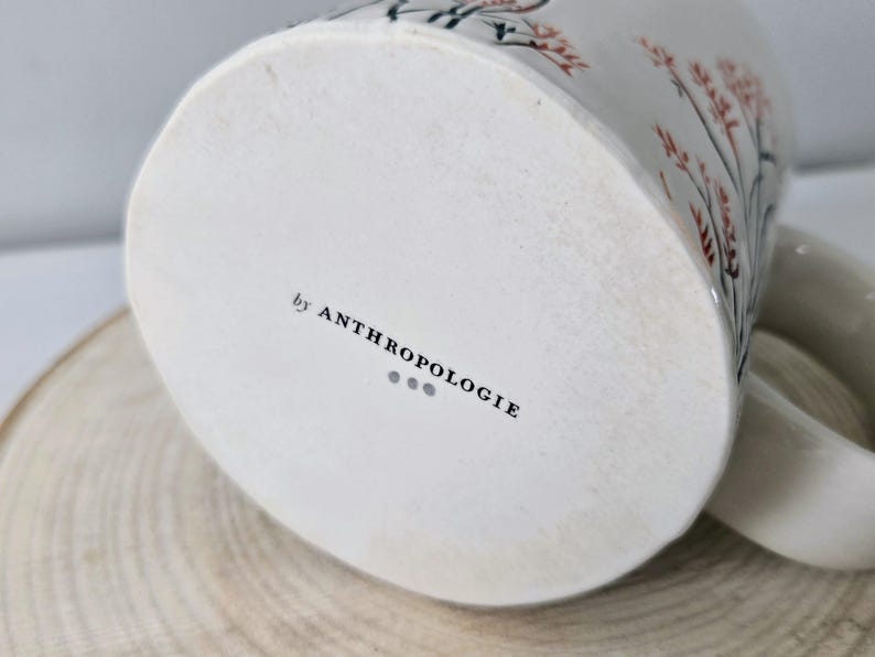 K&ouml;nnte beinhalten: Wei&szlig;e Keramiktasse mit floralem Design in Rot und Schwarz. Die Tasse hat einen gebogenen Henkel und den Text "by ANTHROPOLOGIE" auf der Unterseite. Die Tasse steht auf einer Holzoberfl&auml;che.