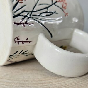 K&ouml;nnte beinhalten: Wei&szlig;e Keramiktasse mit gebogenem Henkel. Die Tasse hat ein Blumendesign mit schwarzen, roten und gr&uuml;nen Akzenten. Die Tasse steht auf einer hellbraunen Holzoberfl&auml;che.
