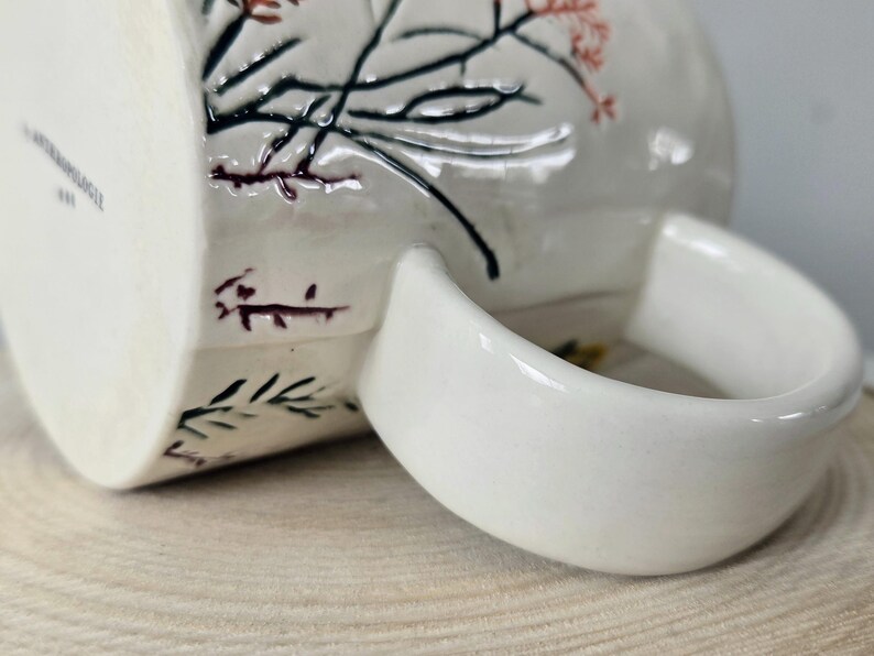 K&ouml;nnte beinhalten: Wei&szlig;e Keramiktasse mit gebogenem Henkel. Die Tasse hat ein Blumendesign mit schwarzen, roten und gr&uuml;nen Akzenten. Die Tasse steht auf einer hellbraunen Holzoberfl&auml;che.