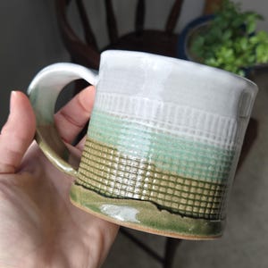 Puede incluir: Una taza de cerámica blanca con un esmalte verde y marrón. La taza tiene un diseño texturizado y una asa ancha.
