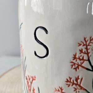 K&ouml;nnte beinhalten: Ein wei&szlig;er Keramikbeh&auml;lter mit einem schwarzen "S" und floralem Design. Der Beh&auml;lter hat orangefarbene und gr&uuml;ne florale Akzente. Das Design ist von Hand auf die Oberfl&auml;che des Beh&auml;lters gemalt.