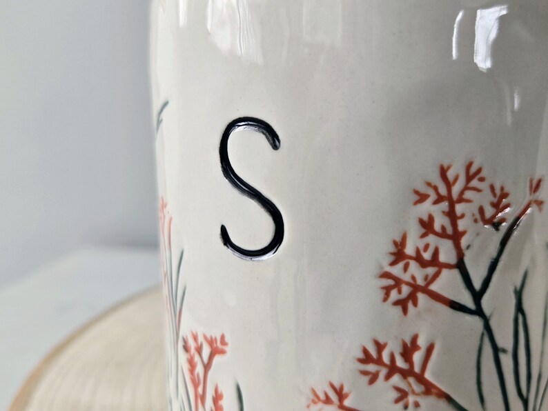 K&ouml;nnte beinhalten: Ein wei&szlig;er Keramikbeh&auml;lter mit einem schwarzen "S" und floralem Design. Der Beh&auml;lter hat orangefarbene und gr&uuml;ne florale Akzente. Das Design ist von Hand auf die Oberfl&auml;che des Beh&auml;lters gemalt.
