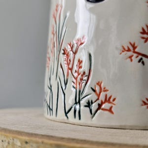 K&ouml;nnte beinhalten: Eine wei&szlig;e Keramik-Tasse mit schwarzem Henkel und dem Text "But First, Coffee" in schwarzer, handschriftlicher Schrift. Die Tasse steht auf einer wei&szlig;en Oberfl&auml;che.
