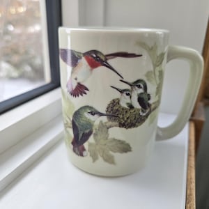 May include: Cream ceramic mug with a hummingbird design. The mug shows a hummingbird in flight, a nest with baby hummingbirds, and a hummingbird perched on a branch. The handle is cream-coloured.