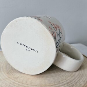 K&ouml;nnte beinhalten: Wei&szlig;er Keramikbecher mit einem floralen Design in Rosa und Schwarz. Der Becher hat einen gebogenen Henkel und den Text "by ANTHROPOLOGIE" auf der Unterseite. Er steht auf einer Holzoberfl&auml;che.
