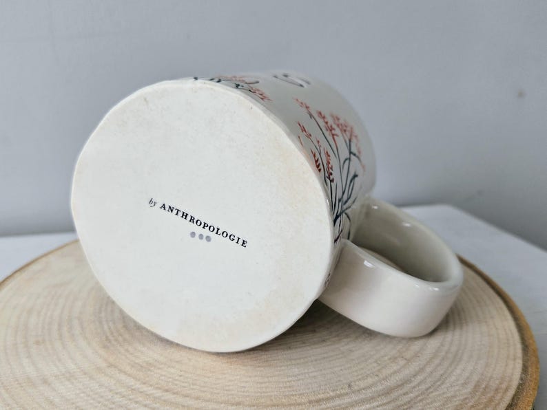 K&ouml;nnte beinhalten: Wei&szlig;er Keramikbecher mit einem floralen Design in Rosa und Schwarz. Der Becher hat einen gebogenen Henkel und den Text "by ANTHROPOLOGIE" auf der Unterseite. Er steht auf einer Holzoberfl&auml;che.