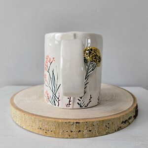 K&ouml;nnte beinhalten: Eine wei&szlig;e Keramiktasse mit gro&szlig;em Henkel, verziert mit handgemalten floralen Designs in Koralle, Schwarz und Gold. Die Tasse wird auf einer runden Holzscheibe pr&auml;sentiert.