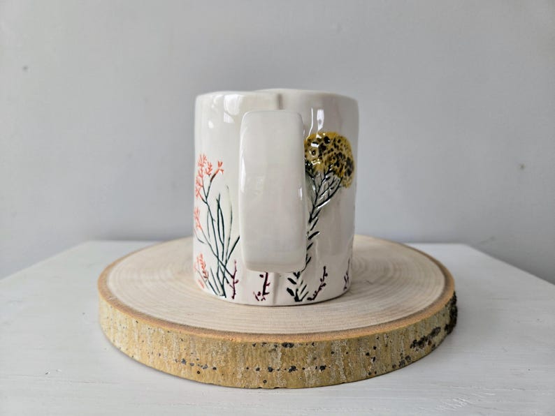 K&ouml;nnte beinhalten: Eine wei&szlig;e Keramiktasse mit gro&szlig;em Henkel, verziert mit handgemalten floralen Designs in Koralle, Schwarz und Gold. Die Tasse wird auf einer runden Holzscheibe pr&auml;sentiert.