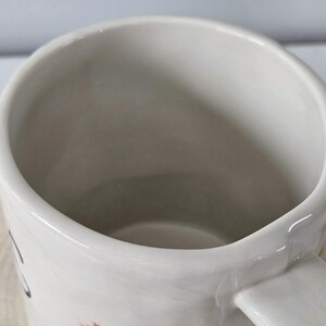 K&ouml;nnte beinhalten: Eine wei&szlig;e Keramiktasse mit gebogenem Henkel. Die Tasse hat eine weite &Ouml;ffnung und eine glatte, gl&auml;nzende Oberfl&auml;che. Das Innere der Tasse ist hellgrau. Die Tasse steht auf einer Holzoberfl&auml;che.