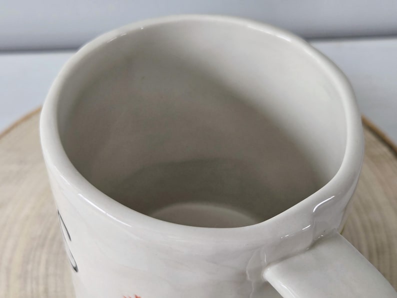 K&ouml;nnte beinhalten: Eine wei&szlig;e Keramiktasse mit gebogenem Henkel. Die Tasse hat eine weite &Ouml;ffnung und eine glatte, gl&auml;nzende Oberfl&auml;che. Das Innere der Tasse ist hellgrau. Die Tasse steht auf einer Holzoberfl&auml;che.