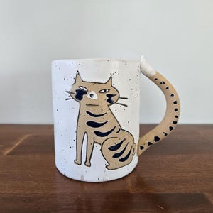 Puede incluir: Taza de cerámica blanca con un diseño de gato pintado a mano. El gato es de color marrón claro con rayas negras y una cola a juego que forma el asa. La taza tiene un acabado moteado y está sobre una superficie de madera.
