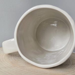 K&ouml;nnte beinhalten: Eine wei&szlig;e Keramiktasse mit abgerundetem Rand und Henkel. Die Tasse steht auf einer Holzoberfl&auml;che, das Innere der Tasse ist sichtbar. Die Tasse hat eine einfarbige, cremewei&szlig;e Farbe.