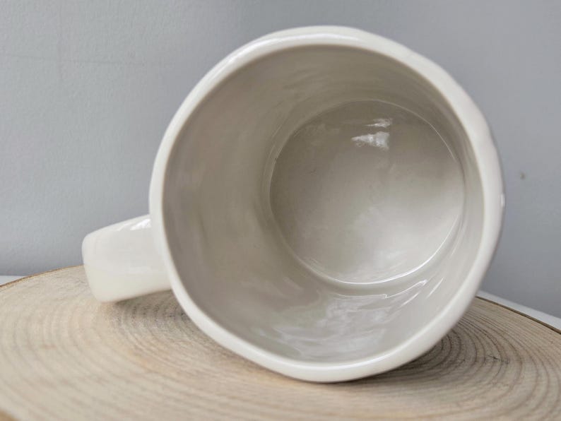 K&ouml;nnte beinhalten: Eine wei&szlig;e Keramiktasse mit abgerundetem Rand und Henkel. Die Tasse steht auf einer Holzoberfl&auml;che, das Innere der Tasse ist sichtbar. Die Tasse hat eine einfarbige, cremewei&szlig;e Farbe.
