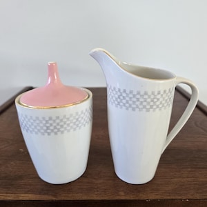 May include: A white ceramic creamer and sugar bowl set. The sugar bowl has a pink lid and gold trim. Both pieces feature a grey checkered pattern around the middle. The creamer has a handle and a spout. The set is on a wooden surface.