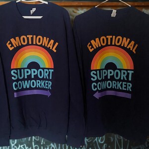 May include: Two navy blue sweatshirts with the text "EMOTIONAL SUPPORT COWORKER" above a rainbow graphic. The text and rainbow are in orange, yellow, green, blue, and purple. A purple arrow points left.